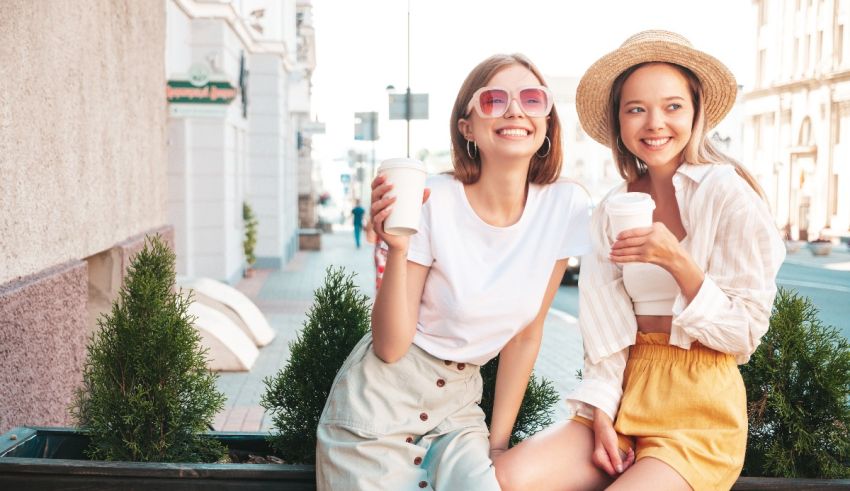 Two young women sitting on a bench and drinking coffee.