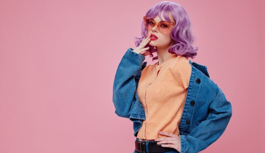 A young woman with purple hair wearing a denim jacket and sunglasses on a pink background.