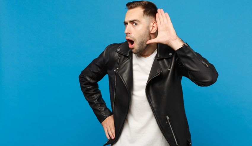 A man in a leather jacket covering his ears with his hand on a blue background.