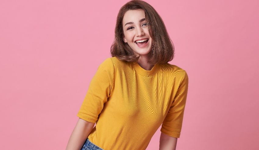 A smiling young woman in a yellow top and jeans posing on a pink background.