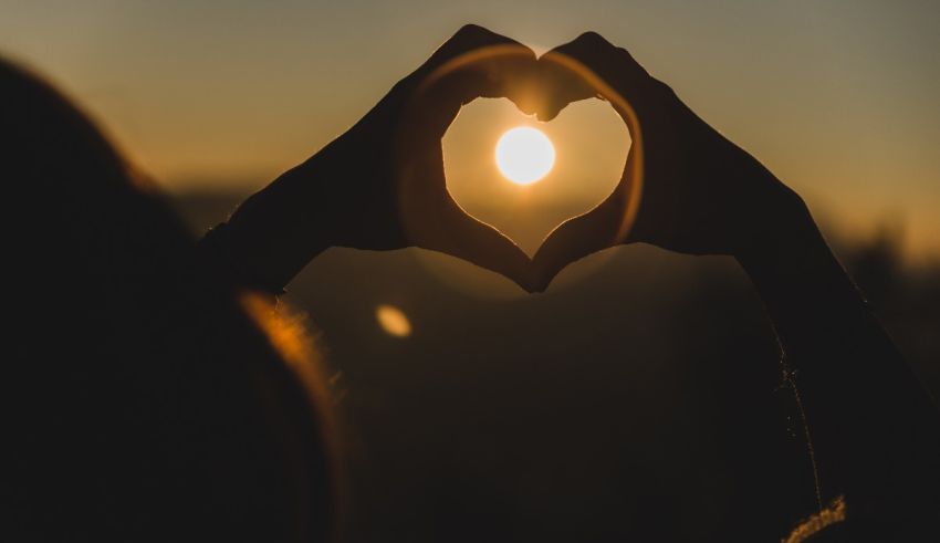 A woman's hands make a heart shape with the sun behind them.