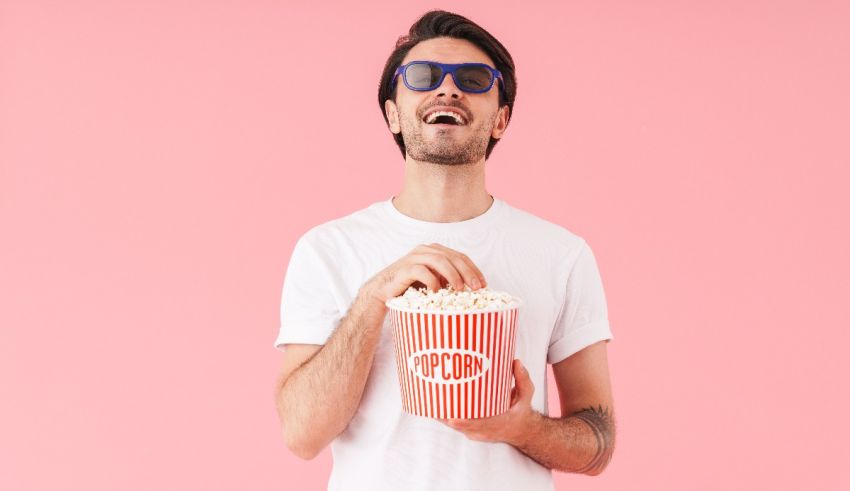 A man wearing sunglasses holding a popcorn box on a pink background.