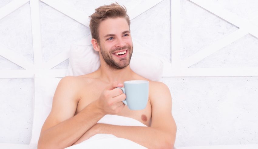 A shirtless man laying in bed with a cup of coffee.