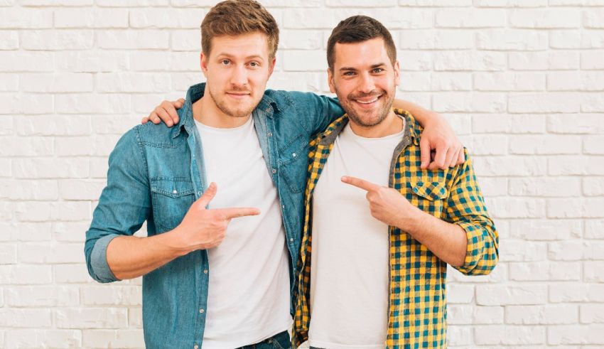 Two young men pointing at each other in front of a white brick wall.