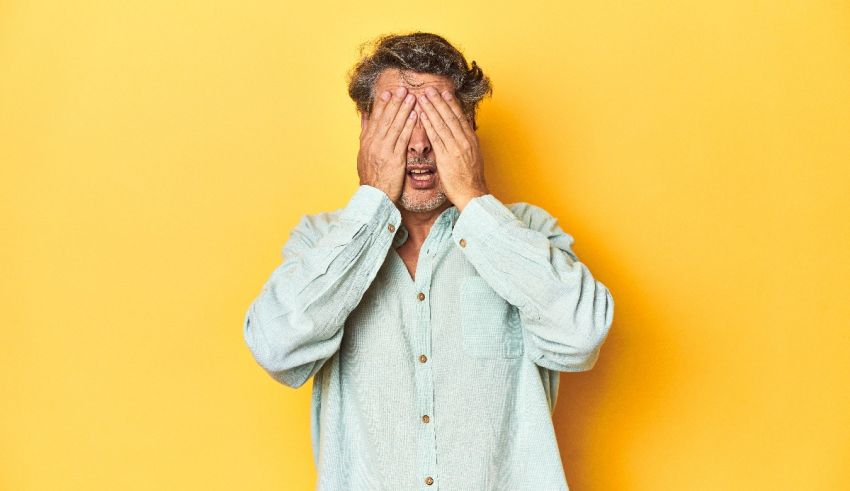 A man covering his eyes with his hands on a yellow background.