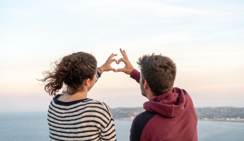 A couple making a heart shape with their hands while looking at the ocean.