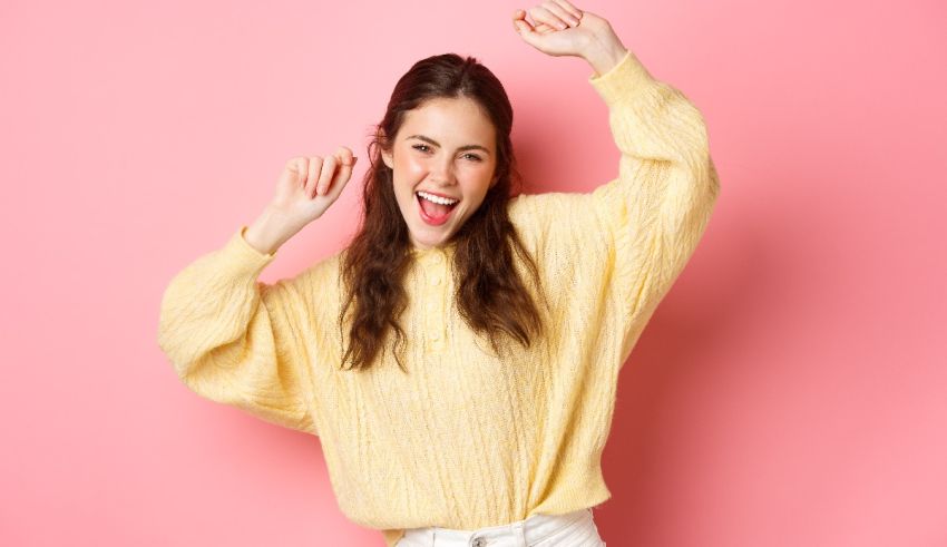 A young woman in a yellow sweater with her arms raised over a pink background.