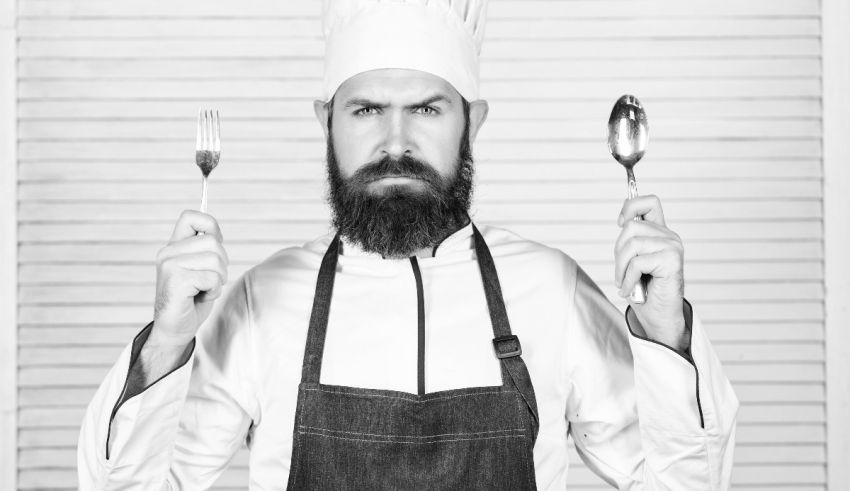 A bearded chef holding a fork and spoon.