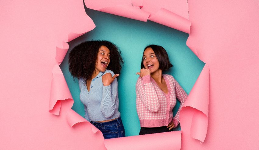 Two women laughing through a hole in pink paper.