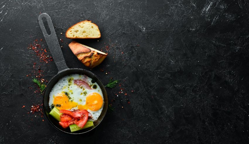 A frying pan with eggs, tomatoes and bread on a dark background.