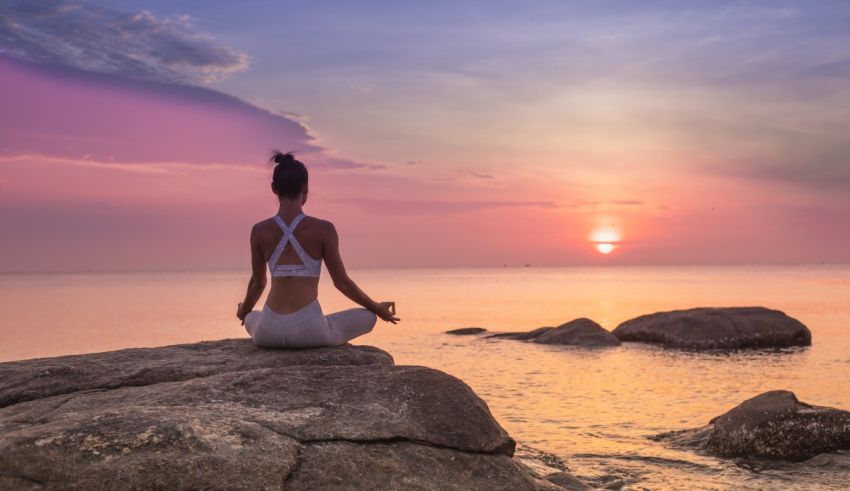 A woman is sitting on rocks and meditating at sunset.