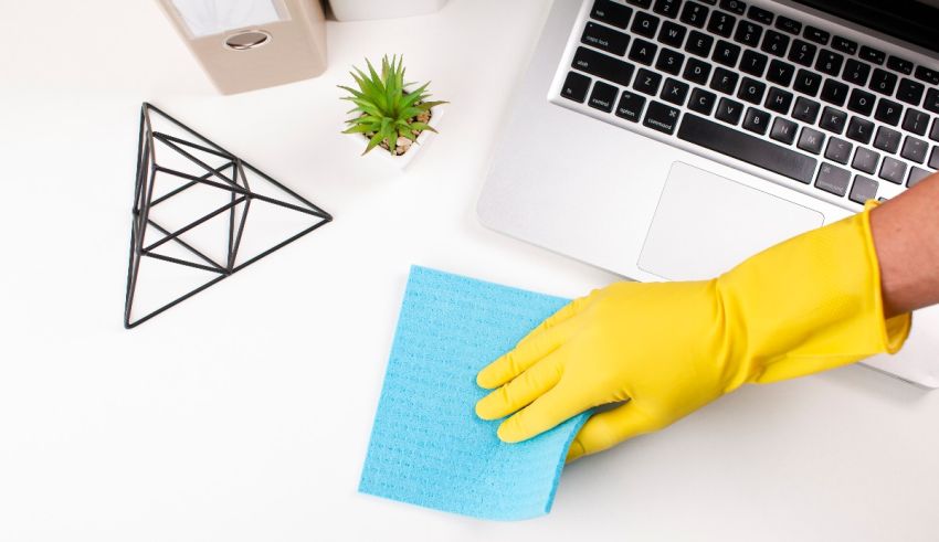 A person wearing a yellow rubber glove cleaning a desk with a laptop.