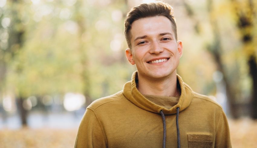 A young man in a yellow sweatshirt is smiling in the park.