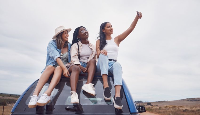 Three young women sitting on the back of a car taking a selfie.