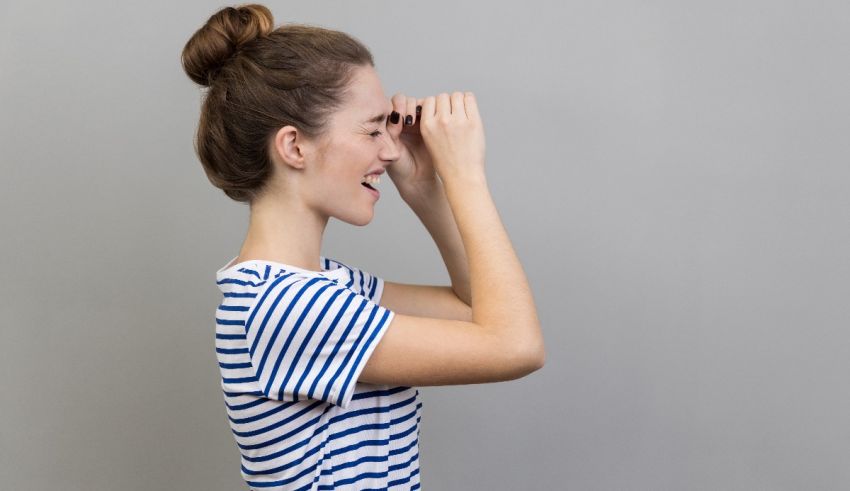 Young woman looking through binoculars on gray background.