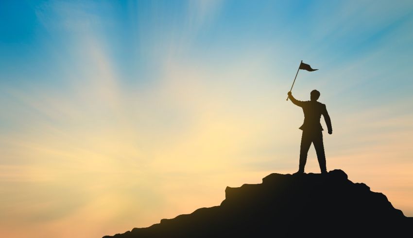 A silhouette of a businessman holding a flag on top of a mountain.