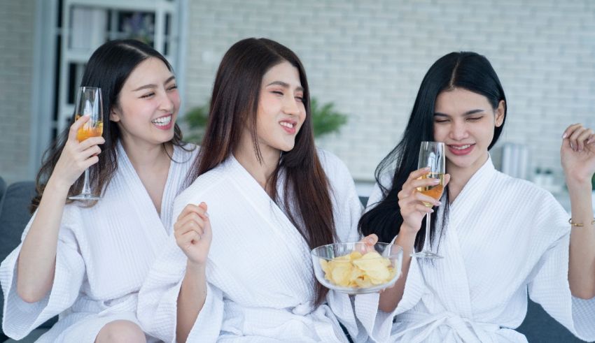 Three asian women in bathrobes sitting on a couch and drinking wine.