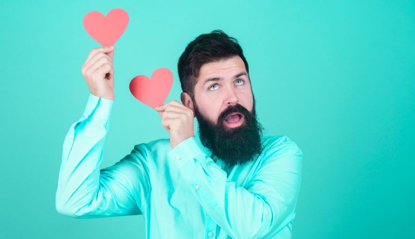 A bearded man with a beard holding heart shaped paper over a turquoise background.