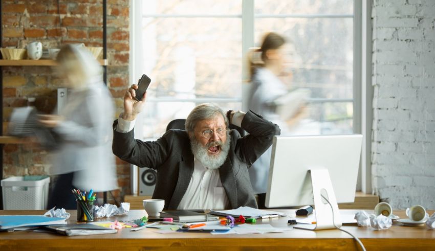 A man with a beard is sitting at a desk with other people around him.