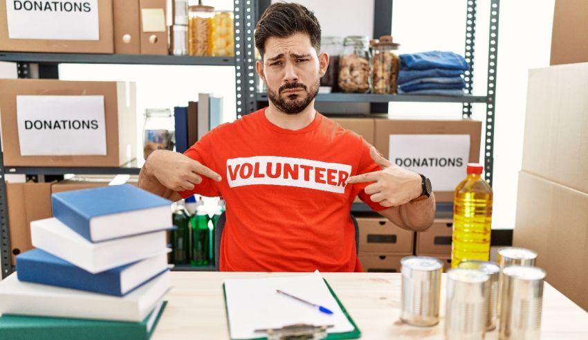 A man in a red t - shirt is pointing at a stack of books.