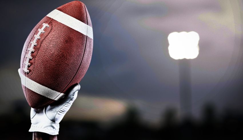 A hand holding an american football in front of a stadium.