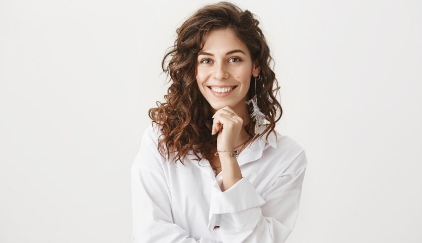 A young woman in a white shirt is posing with her hand on her chin.