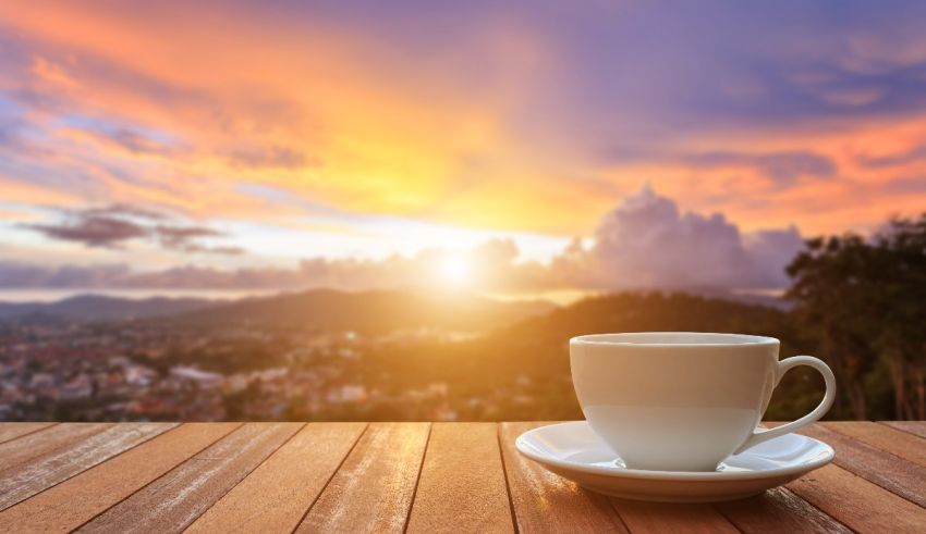 A cup of coffee on a wooden table with a view of a city at sunset.