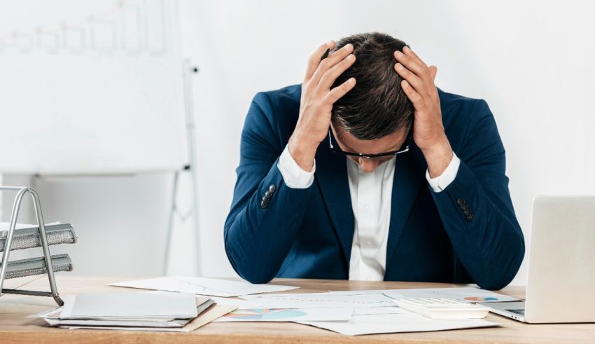 A man in a suit is holding his head while sitting at a desk.