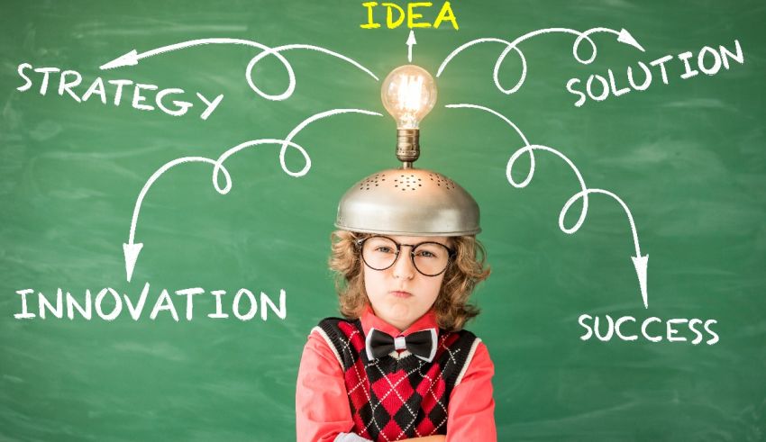 A young boy standing in front of a chalkboard with an idea on his head.