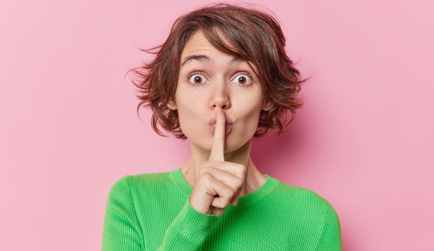 A woman is making a hush sign with her finger on a pink background.